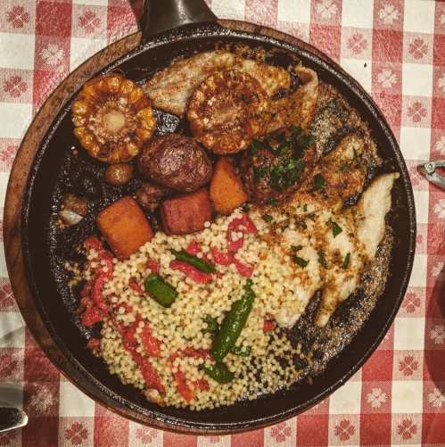 A sizzling platter of grilled fish, vegetables, and couscous, served on a checkered tablecloth.
