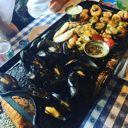 A seafood platter featuring shrimp, crab, mussels, and a dipping sauce, served on a black tray with bread on the side.