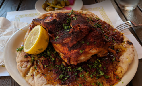 A plate of spiced chicken on flatbread, garnished with herbs and a lemon wedge, served with pickled vegetables.