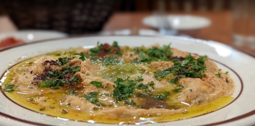 A plate of hummus drizzled with olive oil and garnished with fresh herbs, served in a restaurant setting.