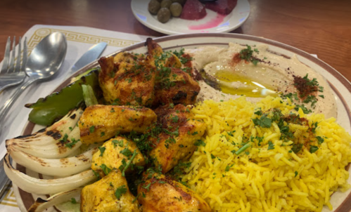 A plate of yellow rice, grilled chicken, hummus with olive oil, and vegetables, served with a side of olives and fruit.