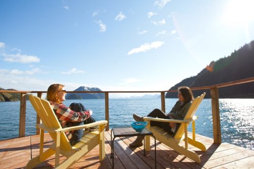 Two people relax in yellow chairs on a deck by the water, surrounded by mountains and a clear blue sky.