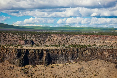 A vast landscape featuring a rugged canyon, layered cliffs, and distant mountains under a cloudy sky.