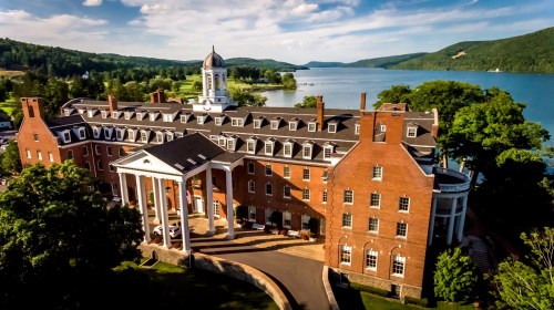 Aerial view of a large brick building with columns, surrounded by trees and a lake, under a blue sky with clouds.