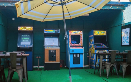 A colorful arcade with vintage game machines under a yellow umbrella, surrounded by tables and chairs on green turf.