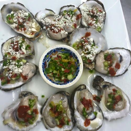 A platter of fresh oysters topped with sauce, cheese, and chopped vegetables, served with a small bowl of dipping sauce.