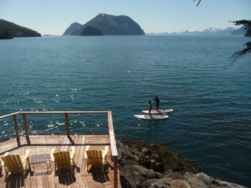 Two people paddleboarding on calm water near a wooden deck with chairs, surrounded by mountains and clear blue skies.