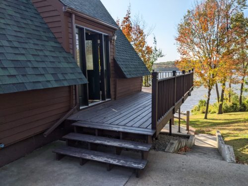 A wooden deck leads to a brown house by a lake, surrounded by trees with autumn foliage.