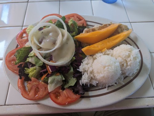 A plate with a fresh salad, sliced tomatoes, rice, and mango slices, served with a creamy dressing.