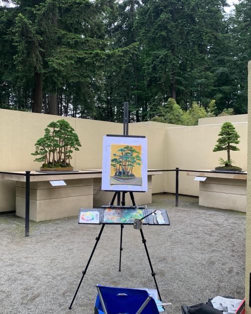 An outdoor art space featuring two bonsai trees and an easel with a painting of trees against a forest backdrop.