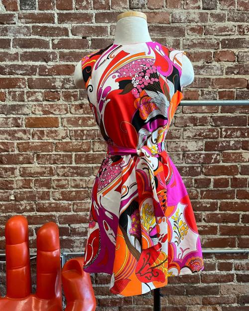 A vibrant, patterned dress on a mannequin, featuring bold colors and a tied waist, against a brick wall backdrop.