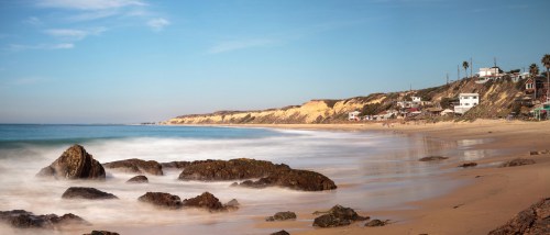 A serene beach scene with smooth waves, rocky shore, and coastal cliffs under a clear blue sky.