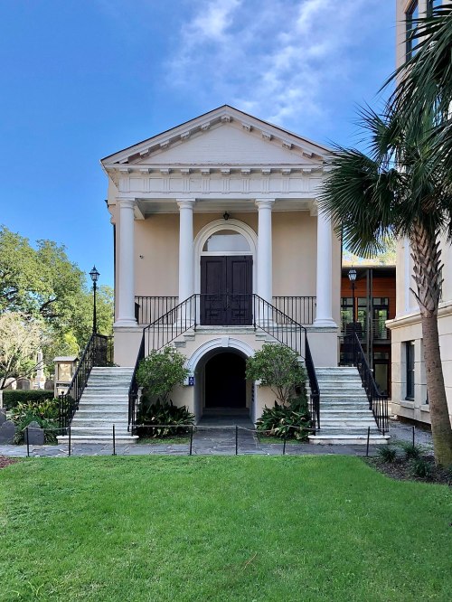 Historic building with a grand entrance, featuring columns, stairs, and lush greenery in front under a blue sky.