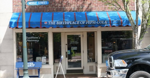 Exterior of a building with a blue awning reading "The Birthplace of Pepsi-Cola" and a pharmacy sign.
