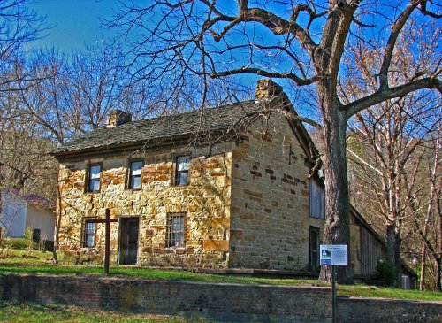 Historic stone house surrounded by trees, with a clear blue sky in the background. Sign visible in front.