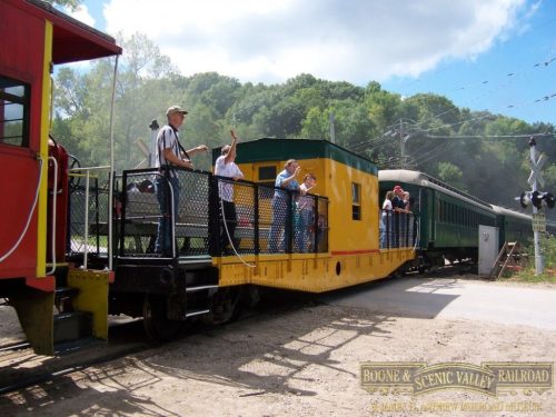 A yellow train car with passengers waving, alongside a red train car, at a scenic railroad crossing.