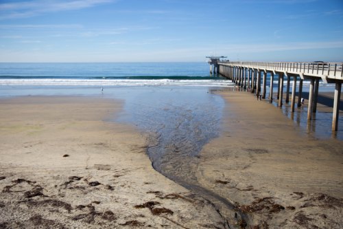 A serene beach scene with gentle waves, a sandy shore, and a long pier extending into the calm ocean under a clear blue sky.