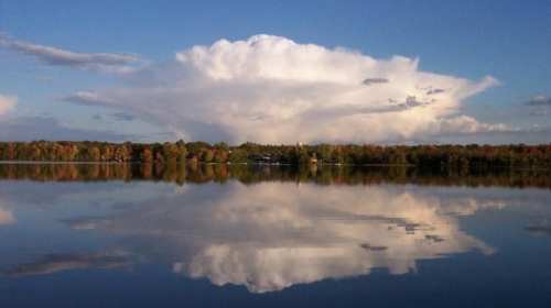A serene lake reflects a large, fluffy cloud against a blue sky, surrounded by colorful autumn trees.