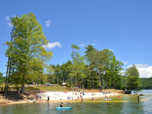 A sunny lakeside scene with people swimming, kayaking, and relaxing on a sandy beach surrounded by trees.