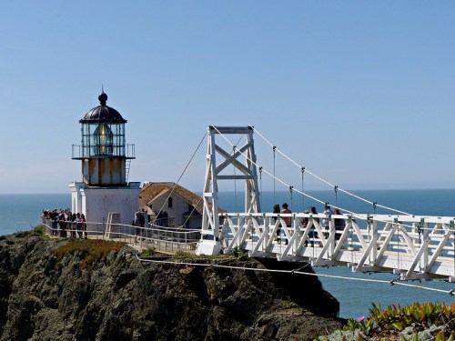 A lighthouse on a rocky shore, connected by a white bridge, with people walking and a clear blue sky above.