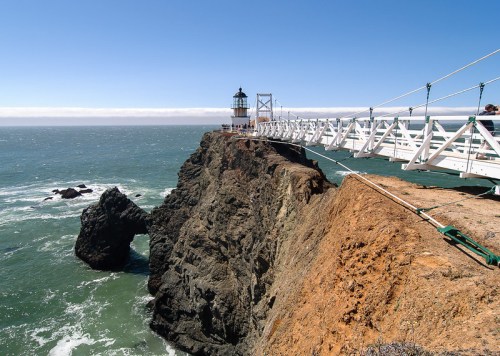 A lighthouse on a rocky cliff, connected by a white bridge, overlooking the ocean under a clear blue sky.