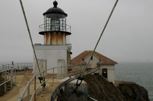 A lighthouse on a rocky shore, surrounded by fog, with a small building nearby and cables extending across the path.