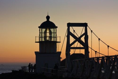 Silhouette of a lighthouse at sunset, with a bridge and people in the foreground against a colorful sky.
