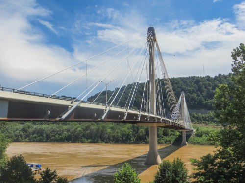 A cable-stayed bridge spans a river, surrounded by green hills and a partly cloudy sky.