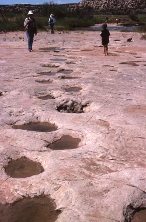 Two people walk on a rocky surface with visible footprints and shallow water pools in a natural landscape.