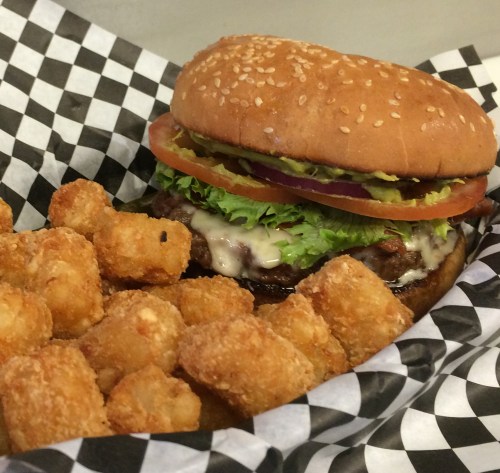 A sesame seed burger with lettuce, tomato, onion, and pickles, served with crispy tater tots in a checkered basket.