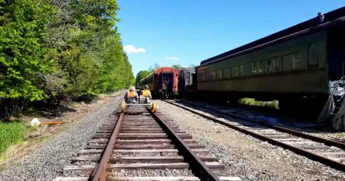 Two people ride a rail cart along train tracks beside vintage train cars and lush greenery under a clear blue sky.