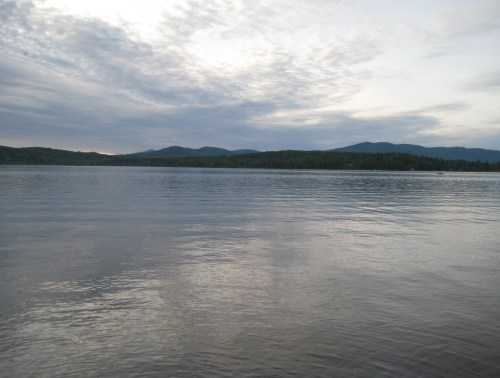 A calm lake reflects the sky, surrounded by mountains and trees under a cloudy sunset.