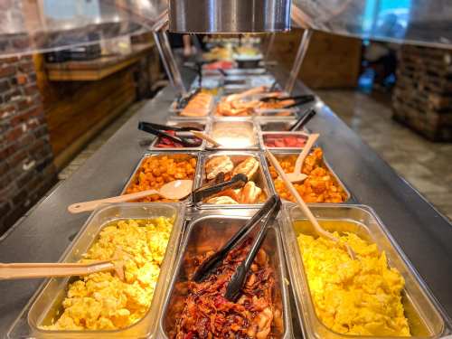 A buffet table featuring various dishes, including scrambled eggs, bacon, and sweet potatoes, with serving utensils.