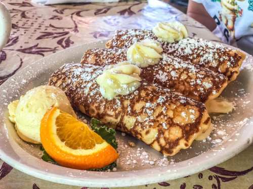 Three crepes filled with cream, topped with powdered sugar and butter, served with a scoop of ice cream and an orange slice.