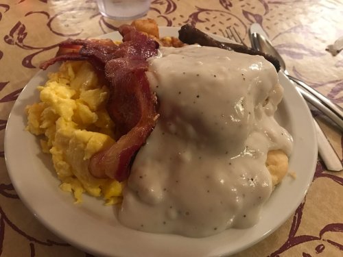 A plate of breakfast featuring scrambled eggs, bacon, sausage, and biscuits smothered in creamy gravy.