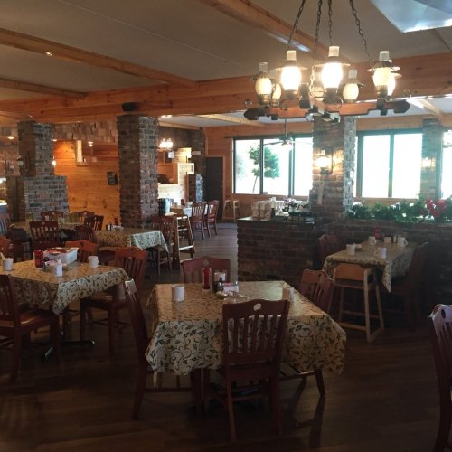 Cozy restaurant interior with wooden beams, stone walls, and tables set with floral tablecloths. Warm lighting from chandeliers.