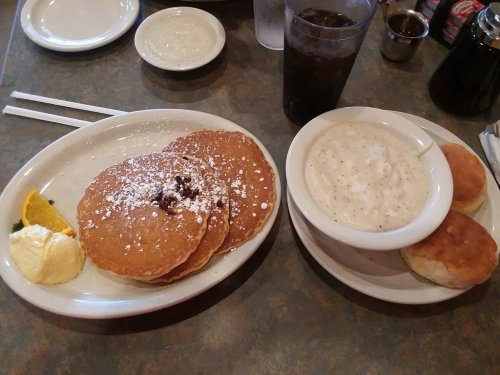 A plate of pancakes with butter and powdered sugar, served with a bowl of gravy and biscuits, alongside a drink.