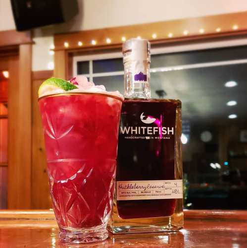 A vibrant red cocktail with ice and a lime wedge next to a bottle of Whitefish blueberry liqueur on a bar.