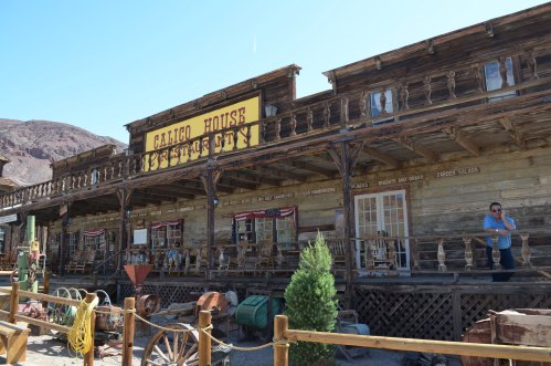 A rustic wooden building with a sign reading "Calico House" in a desert setting, surrounded by vintage items.