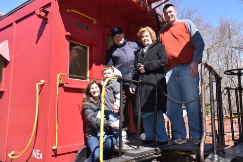 A family poses together on a red train car, smiling and enjoying a sunny day outdoors.