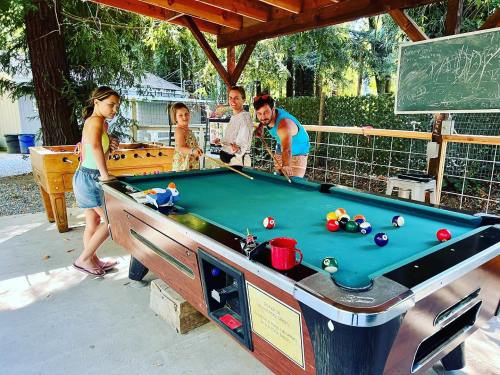 A group of four people playing pool outdoors under a wooden shelter, surrounded by trees and a chalkboard.