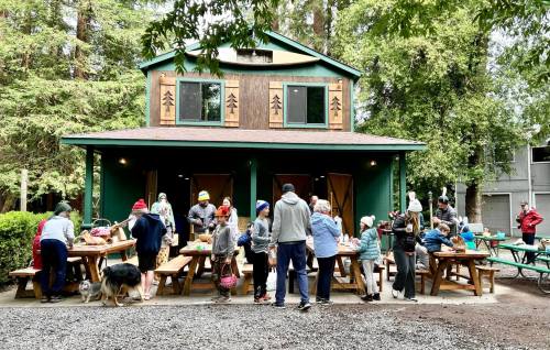 A group of people gather outside a green building, enjoying activities at picnic tables surrounded by trees.