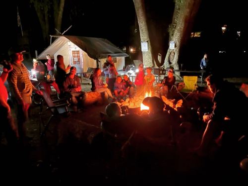A group of people gathered around a campfire at night, with tents and trees in the background.