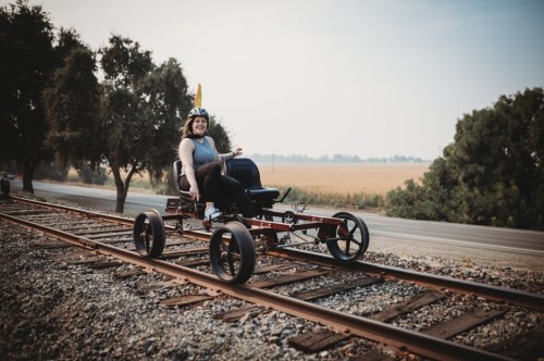 A person in a helmet sits on a go-kart on railway tracks, surrounded by trees and fields in the background.