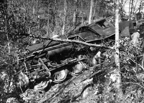 A derailed train car rests on its side among trees and brush, with visible wheels and debris scattered around.