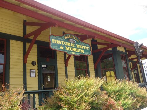 Sign for Saluda Historic Depot & Museum on a yellow building with red accents and surrounding greenery.