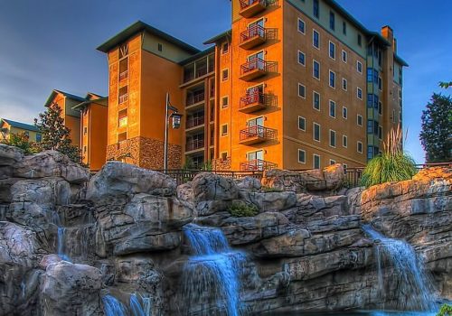 A multi-story building with balconies, surrounded by rocks and a small waterfall, under a clear blue sky.