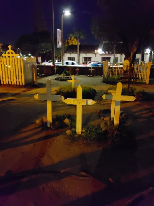 Three white crosses in a dimly lit cemetery at night, surrounded by small plants and a white picket fence.