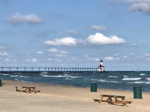 A sandy beach with picnic tables, a pier extending into the water, and a lighthouse under a partly cloudy sky.