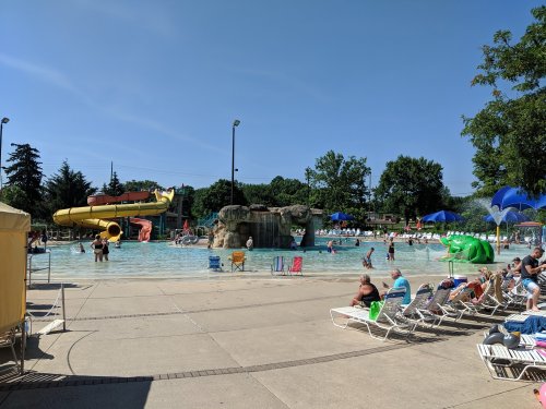 A busy water park with a large pool, water slides, and sunbathers lounging on chairs under umbrellas.